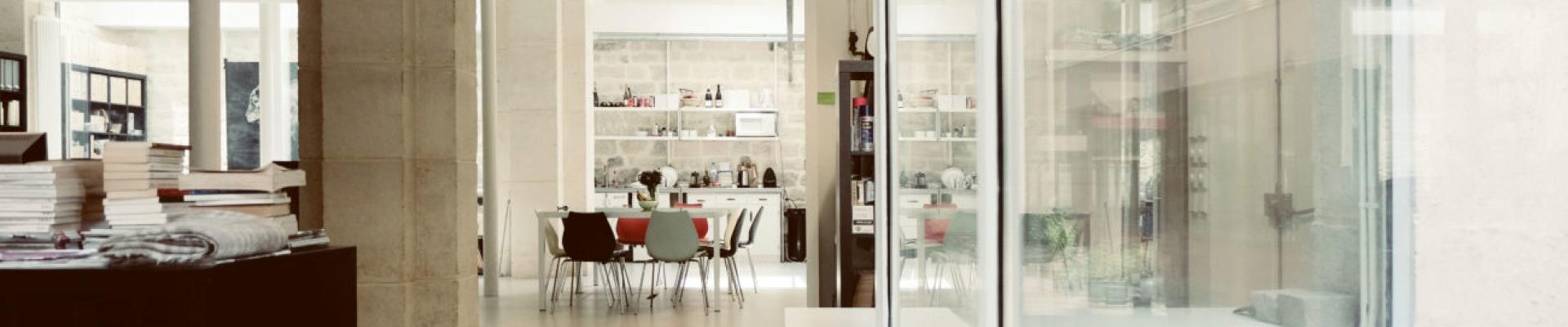 Cette image montre un espace intérieur lumineux et épuré, probablement un bureau ou un atelier de travail. L’architecture mélange des éléments industriels et classiques, avec des colonnes en pierre et des structures métalliques apparentes.